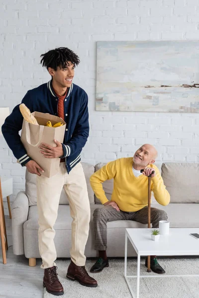 Positive african american grandson holding bag with food near grandpa with walking cane at home — Foto stock