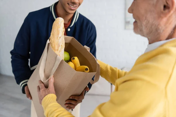Cropped view of blurred senior man taking bag with food from african american grandson at home — Fotografia de Stock