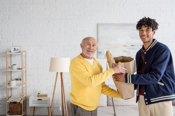 Cheerful senior man and african american grandson holding bag with food at home — Fotografia de Stock