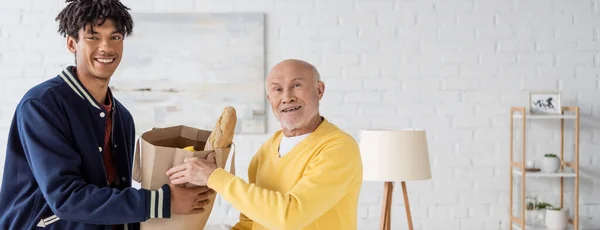 Positive african american man and pensioner holding bag with food at home, banner — Stockfoto