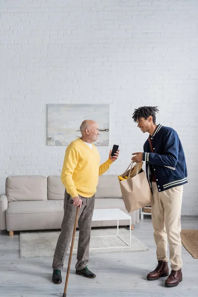 Side view of smiling pensioner holding smartphone near african american grandson with food in bag at home - foto de stock