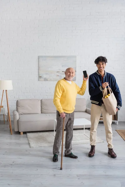 Cheerful pensioner holding smartphone near african american grandson with food at home — Stockfoto
