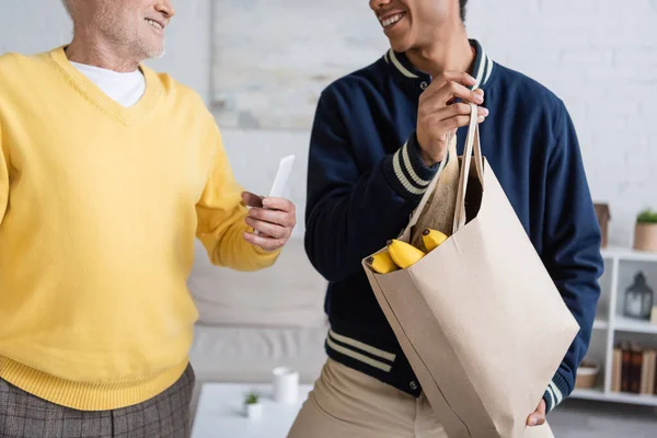 Cropped view of smiling elderly man holding smartphone near african american grandson with food in bag at home — Foto stock