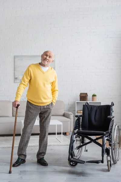 Старший мужчина с тростью, стоящий возле инвалидного кресла дома — стоковое фото