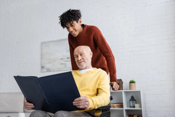 African american man standing near granddad with photo album in wheelchair at home — Foto stock