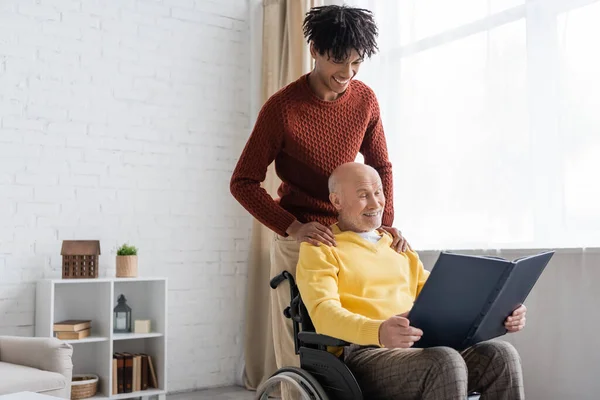 Cheerful african american man hugging granddad with photo album in wheelchair at home — Stockfoto