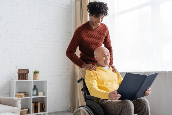 Cheerful african american man hugging granddad with photo album in wheelchair at home — Fotografia de Stock