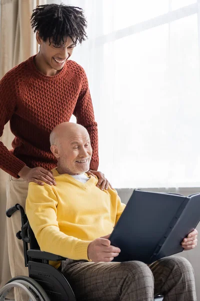 African american grandson hugging grandfather with photo album in wheelchair at home - foto de stock