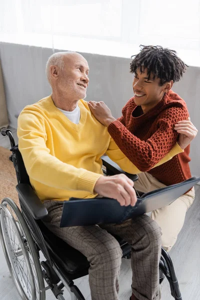 Позитивный африканский внук обнимает дедушку с фотоальбомом в инвалидном кресле дома — стоковое фото