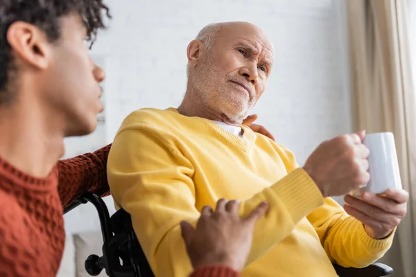 Sad senior man holding cup in wheelchair near blurred african american grandson at home - foto de stock