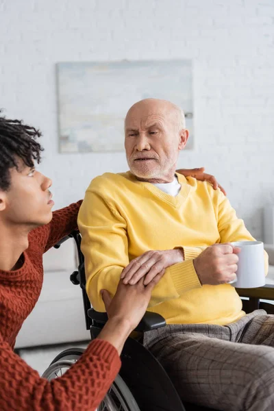 African american grandson touching hand of granddad with cup in wheelchair at home - foto de stock