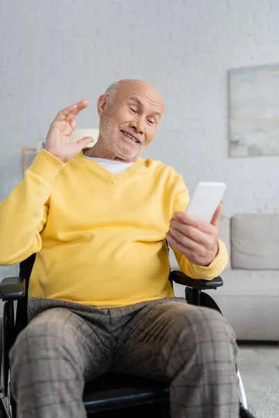 Smiling pensioner having video call on smartphone while sitting in wheelchair at home — Foto stock