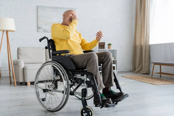 Смеющийся мужчина со смартфоном, сидящий дома в инвалидной коляске — стоковое фото