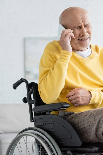 Разочарованный пожилой мужчина разговаривает на смартфоне, сидя дома в инвалидной коляске — стоковое фото
