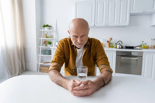 Sad senior man sitting near glass of water in kitchen - foto de stock