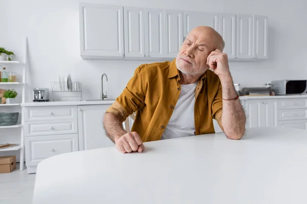 Грустный пожилой человек сидит за белым столом на кухне — стоковое фото