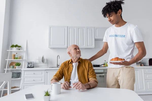 Smiling african american volunteer holding croissant near pensioner at home — Stock Photo