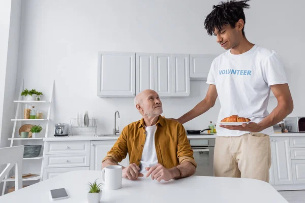 Smiling african american volunteer holding croissant near pensioner at home — Fotografia de Stock