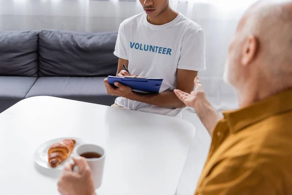 Blurred pensioner pointing with hand near african american volunteer with clipboard at home - foto de stock