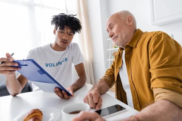 African american volunteer holding clipboard near senior man with cup and smartphone at home — Stock Photo