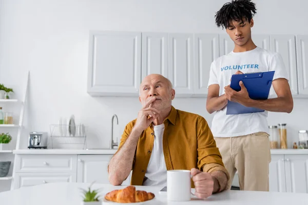Pensive senior man holding cup near croissant and african american volunteer with clipboard at home — Foto stock