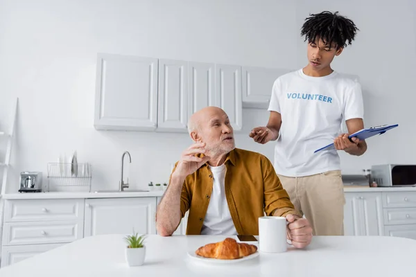 African american volunteer holding clipboard and talking to elderly man with cup at home — Stock Photo
