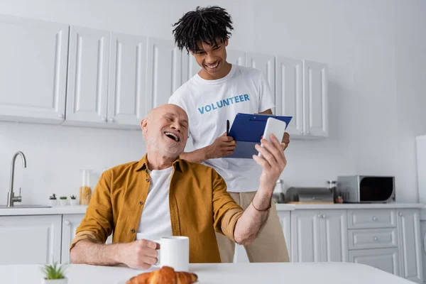 Positive senior man holding cellphone and cup near african american volunteer with clipboard in kitchen — Foto stock