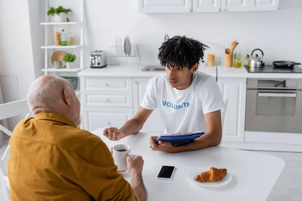 African american volunteer with clipboard talking to senior man with tea in kitchen — Fotografia de Stock