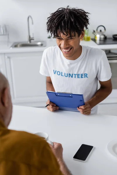 Cheerful african american volunteer with clipboard looking at blurred senior man with tea near smartphone in kitchen - foto de stock