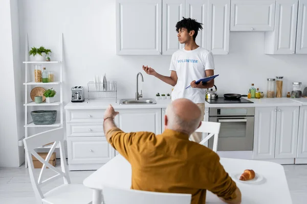 African american volunteer holding clipboard near blurred elderly man pointing with finger in kitchen — Foto stock