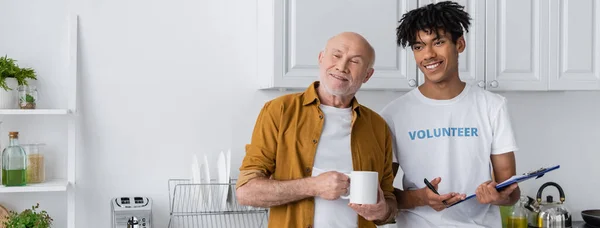 Cheerful african american volunteer holding clipboard near pensioner with cup in kitchen, banner — Foto stock