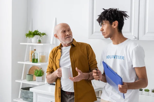 Pensioner holding cup and looking at african american volunteer with clipboard in kitchen — Stock Photo