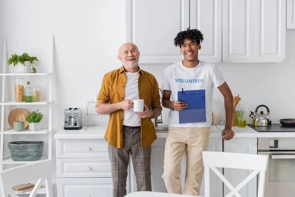 Cheerful african american volunteer with clipboard and elderly man looking at camera in kitchen — Stock Photo