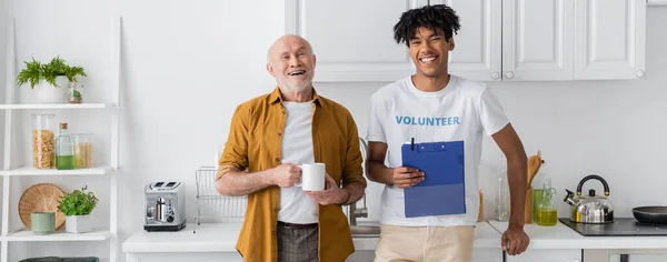 Happy senior man holding cup near african american volunteer with clipboard in kitchen, banner - foto de stock