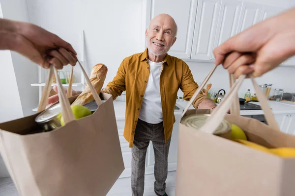 Cheerful pensioner looking at african american volunteer holding bags with food in kitchen - foto de stock