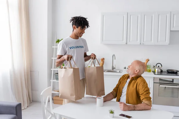 African american volunteer holding bags near elderly man in kitchen - foto de stock