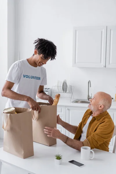Smiling african american volunteer holding package with food near pensioner in kitchen — Stock Photo