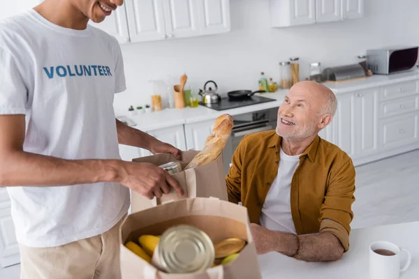 Smiling african american volunteer holding package with food near pensioner in kitchen — Foto stock