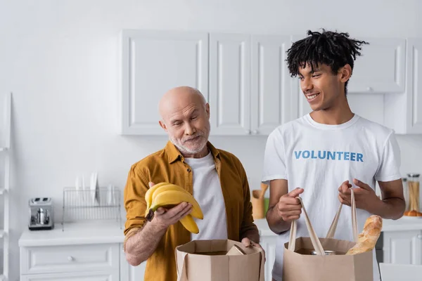 Smiling senior man holding bananas near bags and african american volunteer in kitchen — Foto stock