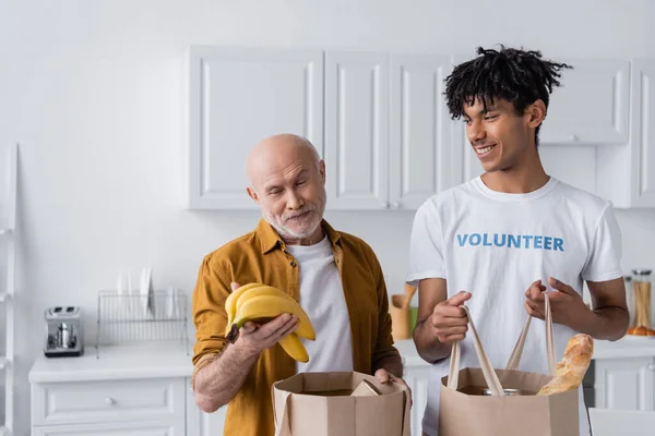 Smiling senior man holding bananas near bags and african american volunteer in kitchen — Fotografia de Stock