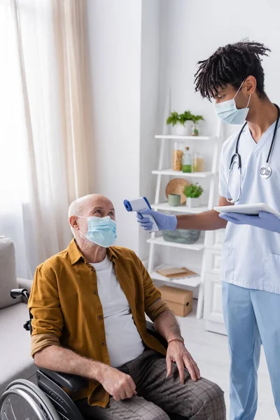 African american nurse with digital tablet holding pyrometer near pensioner in medical mask and wheelchair at home — Stockfoto