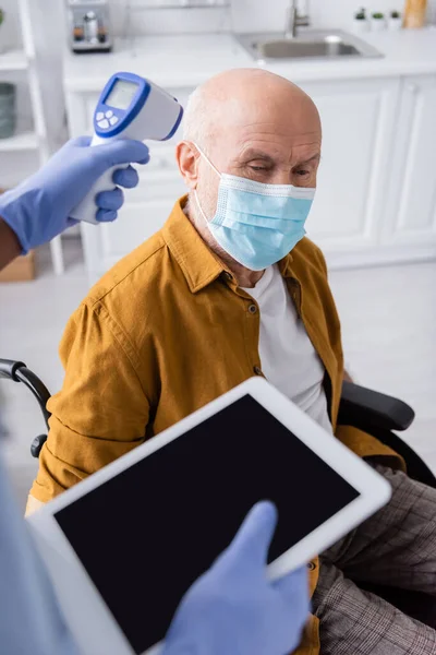 African american nurse holding pyrometer and digital tablet near pensioner in medical mask and wheelchair at home — Fotografia de Stock