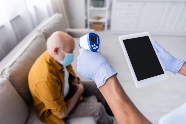African american nurse holding pyrometer and digital tablet with blank screen near blurred pensioner in medical mask at home - foto de stock