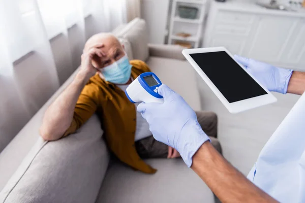 African american nurse holding digital tablet and pyrometer near blurred senior patient in medical mask at home — Foto stock