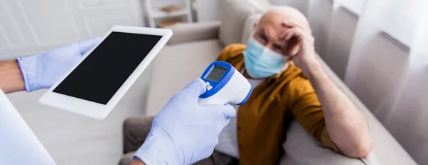 African american nurse in latex gloves holding pyrometer and digital tablet near blurred senior man in medical mask at home, banner - foto de stock