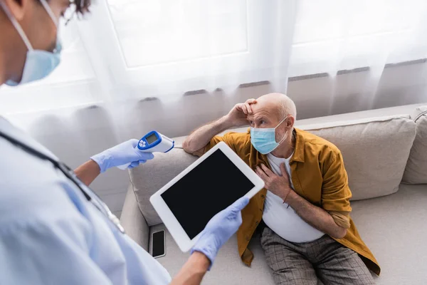 Elderly man in medical mask sitting near blurred african american nurse with pyrometer and digital tablet at home - foto de stock