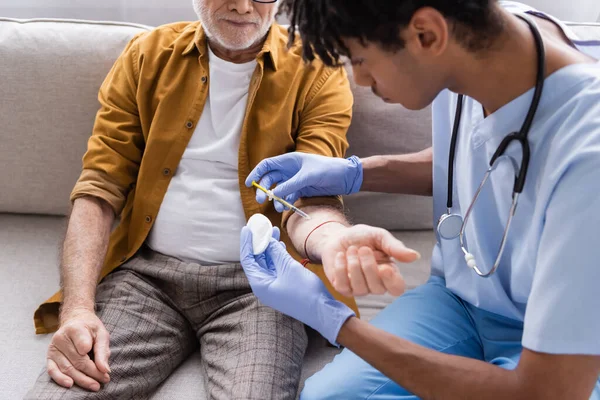 Blurred african american nurse doing injection to pensioner on couch at home - foto de stock