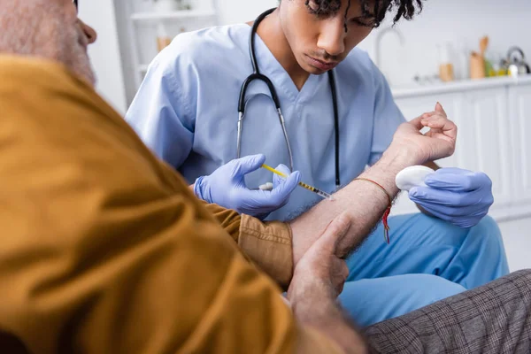 African american nurse in latex gloves and uniform doing injection to elderly patient at home — Stock Photo