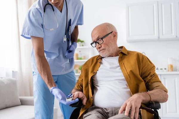 African american nurse with clipboard holding pulse oximeter near patient in wheelchair at home — Foto stock