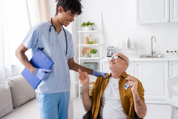 Smiling african american nurse looking at senior patient with pulse oximeter at home — Stock Photo